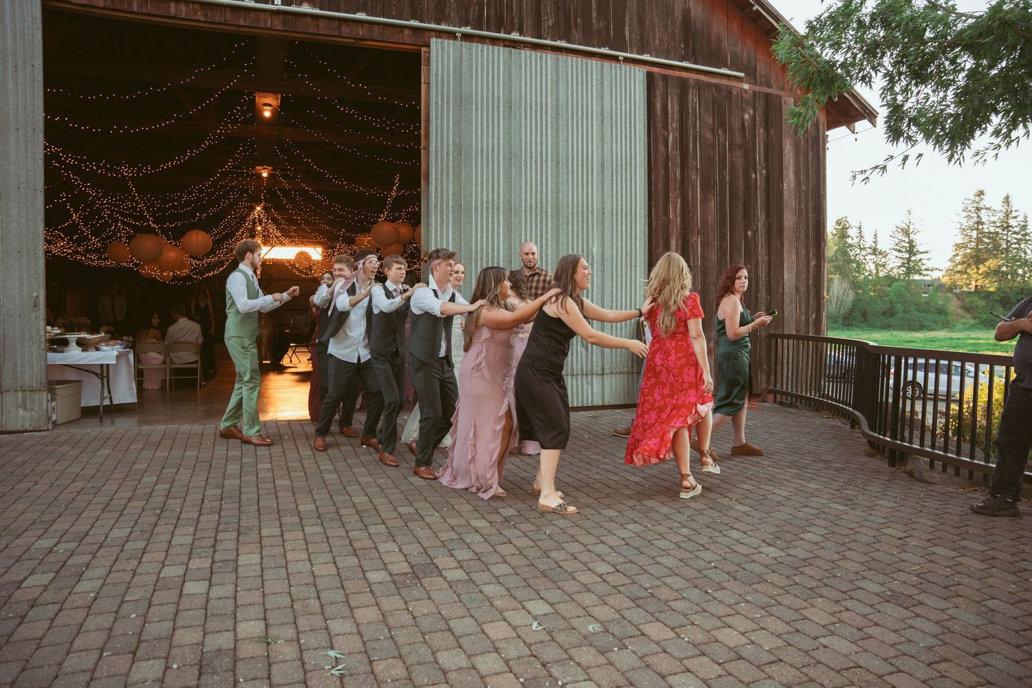 Wedding guests dance in a line formation on a rustic barn patio decorated with string lights at dusk.