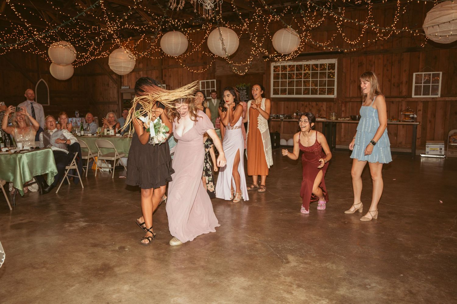 Lively dance floor scene at a barn wedding with string lights and paper lanterns overhead.