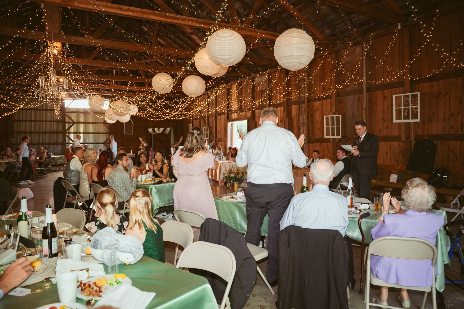 Wedding celebration in a wooden barn with hanging lanterns and guests seated at mint green decorated tables.