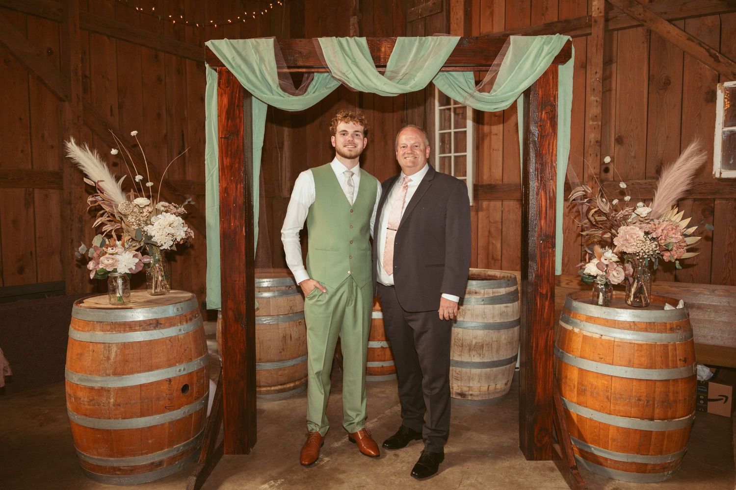 Wedding party members gather between decorative barrels in a barn venue with draped fabric decor.