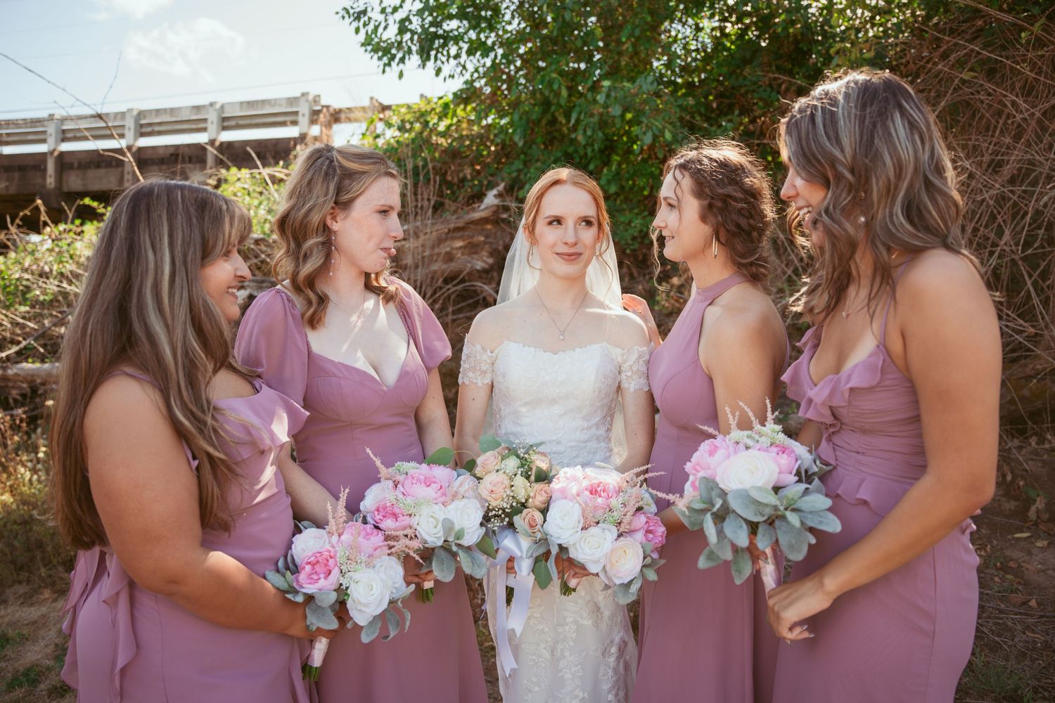 Bridal party in mauve dresses gather for outdoor wedding photos.