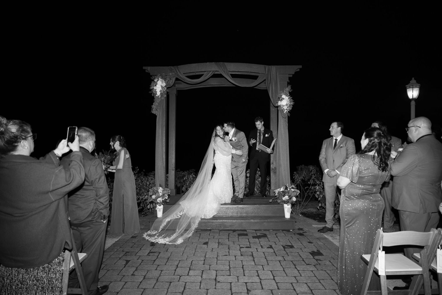 Evening outdoor wedding ceremony with guests seated on wooden chairs facing a decorated arch.