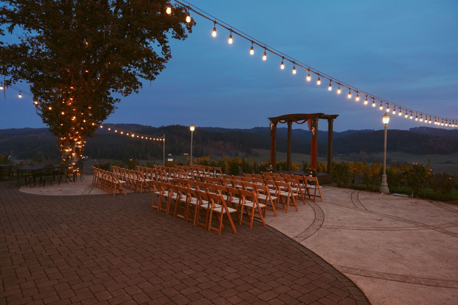 Outdoor wedding ceremony setup with string lights and wooden chairs at dusk.