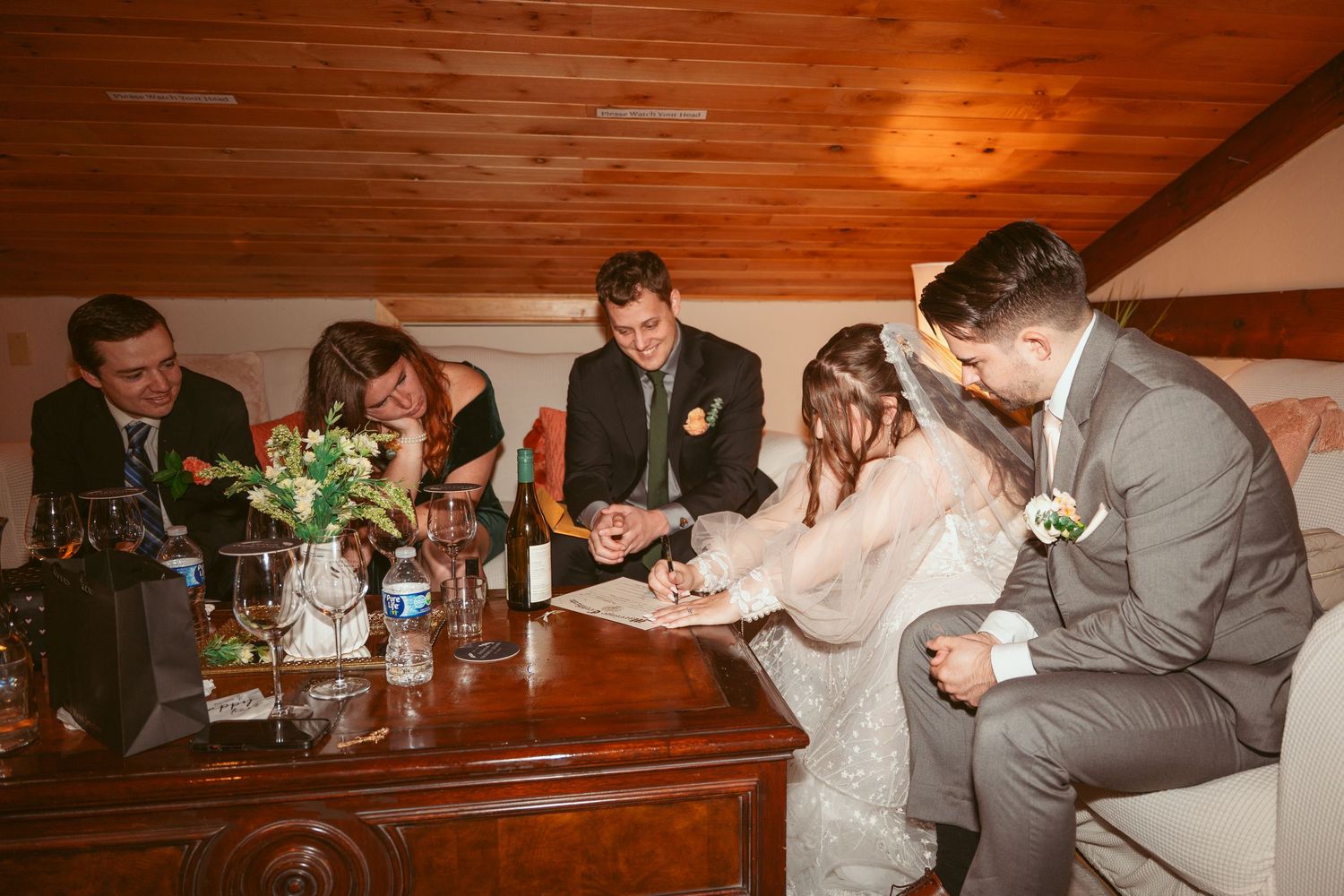 Wedding party members signing official documents at rustic wooden table indoors.