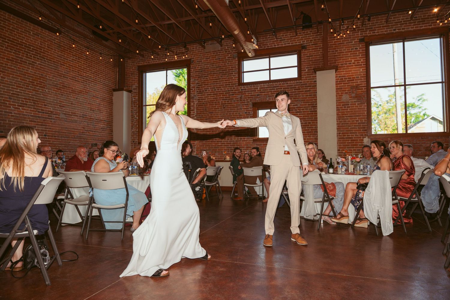 Wedding reception dance in rustic brick building with natural light streaming through windows.