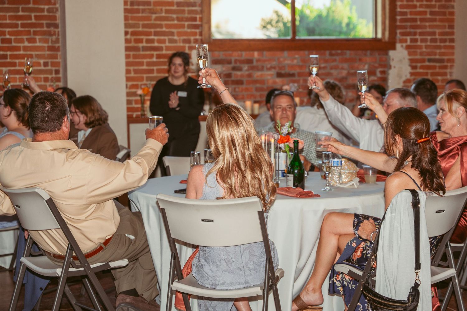 Wedding guests toasting with raised glasses at round tables during the reception celebration.