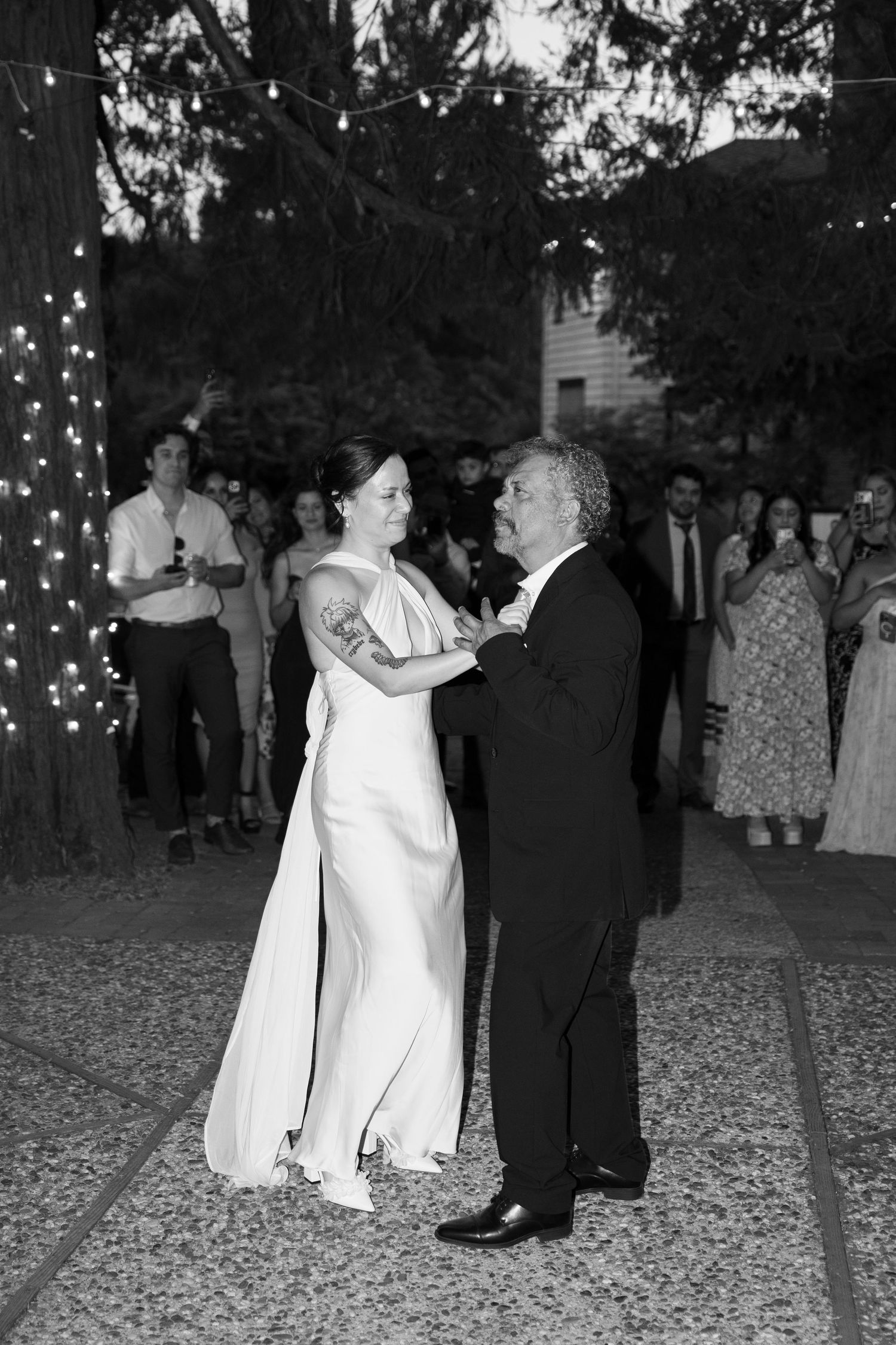 Wedding guests dance together on a gravel dance floor under twinkling lights at an outdoor evening reception.