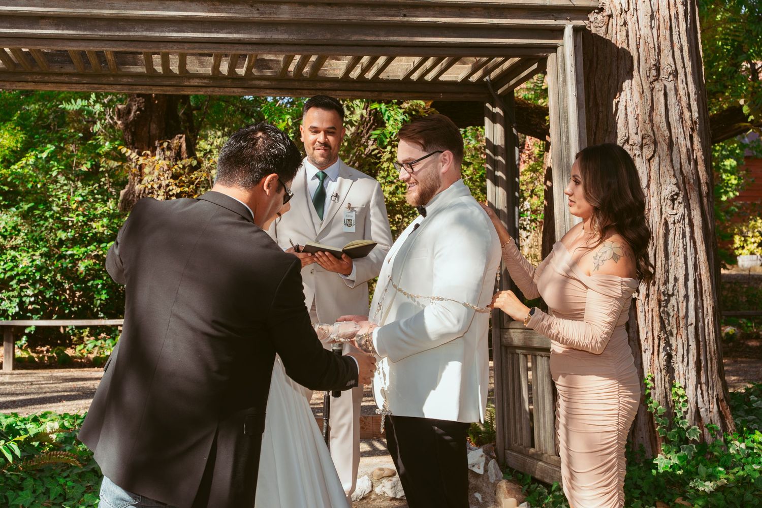 Wedding ceremony taking place under a wooden pergola with guests and officiant gathered around.