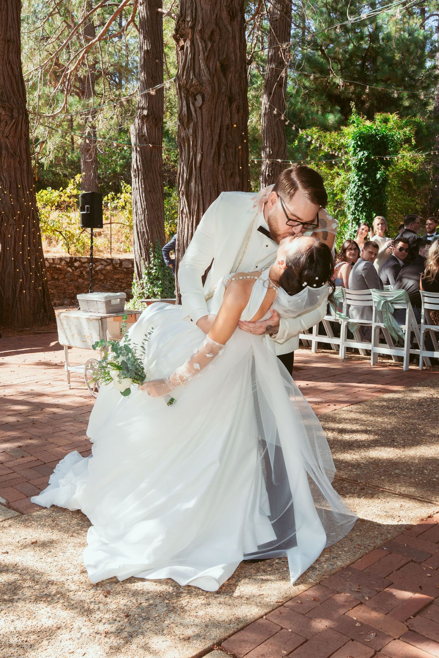 A romantic wedding couple shares a dramatic dip kiss on a brick patio surrounded by tall trees.