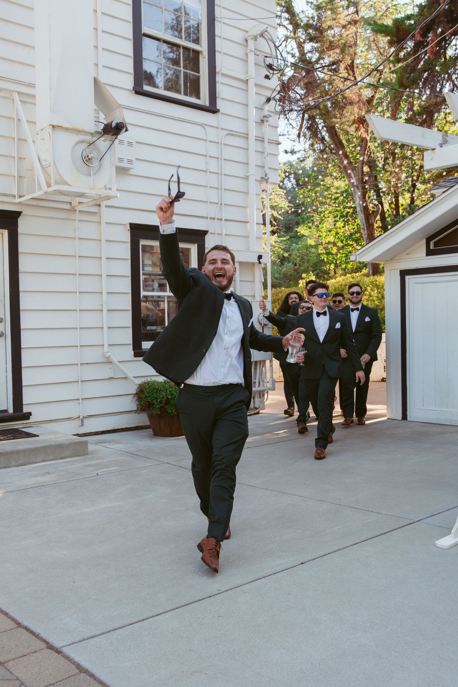 Wedding party members in black tuxedos pose playfully outside a white building on a sunny day.