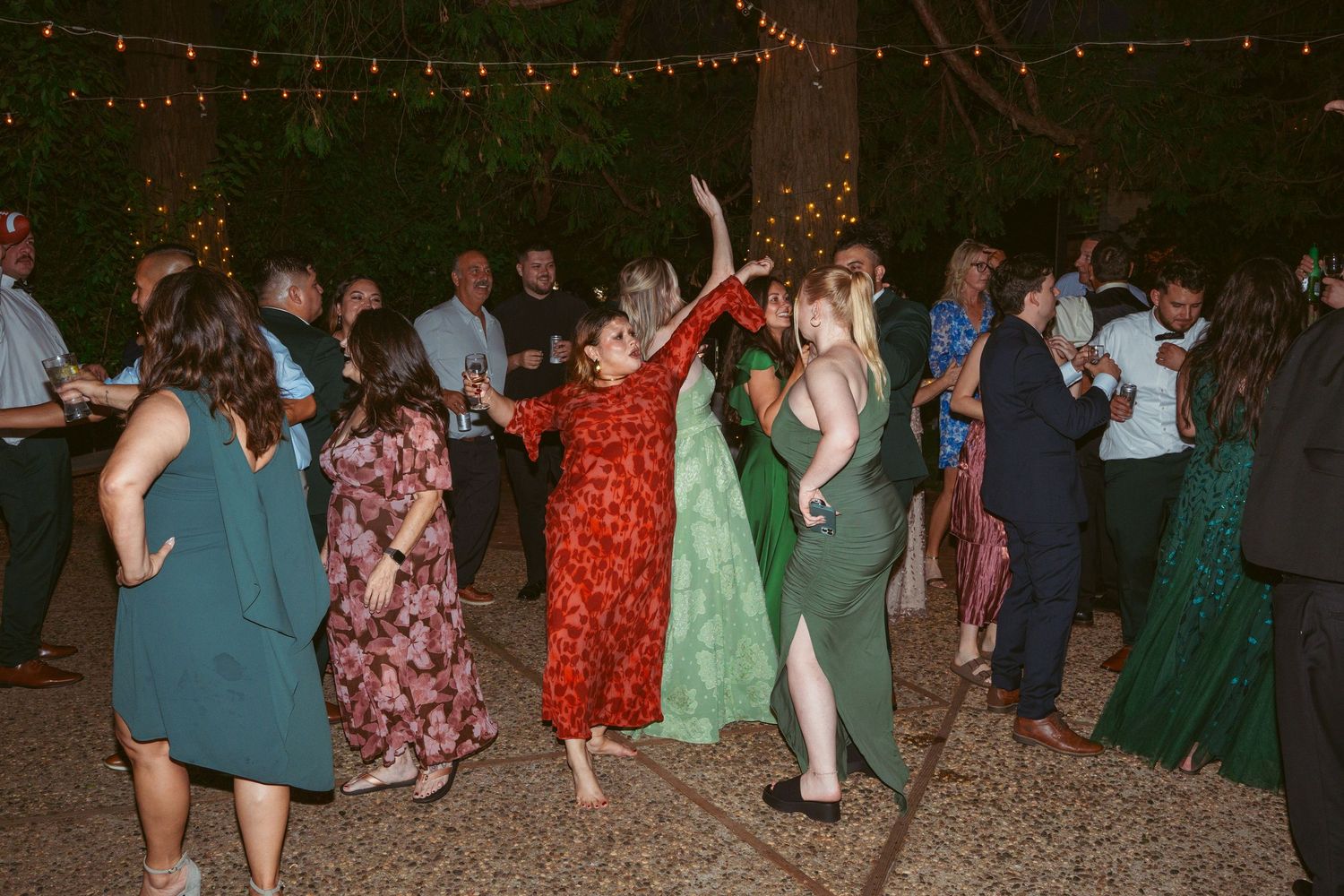 Outdoor wedding reception dance party with guests celebrating under illuminated trees.