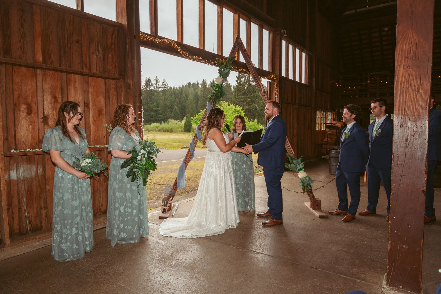 Wedding party poses in rustic barn venue decorated with string lights and wooden beams.