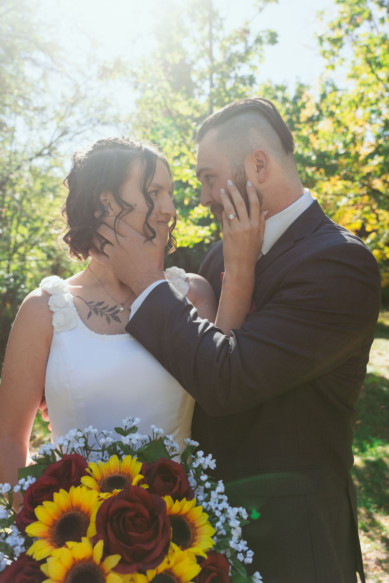 Wedding couple embracing outdoors with sunflower and rose bouquet against natural backdrop.