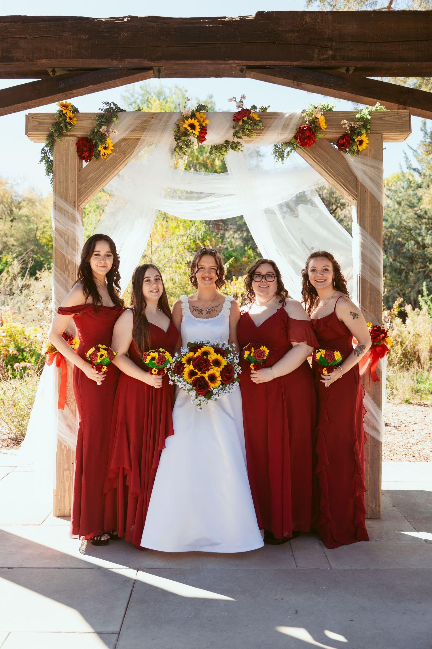 Wedding party with bridesmaids in deep red dresses and sunflower bouquets under wooden pavilion.