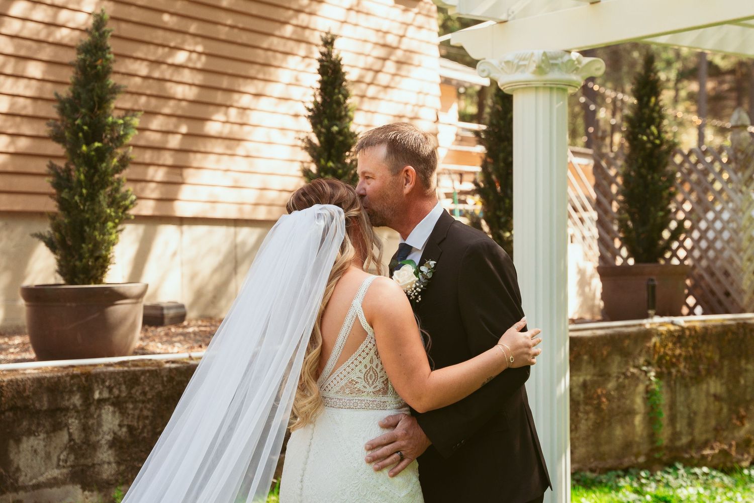 Romantic wedding couple embraces in a sunlit garden setting with white pillars and greenery.