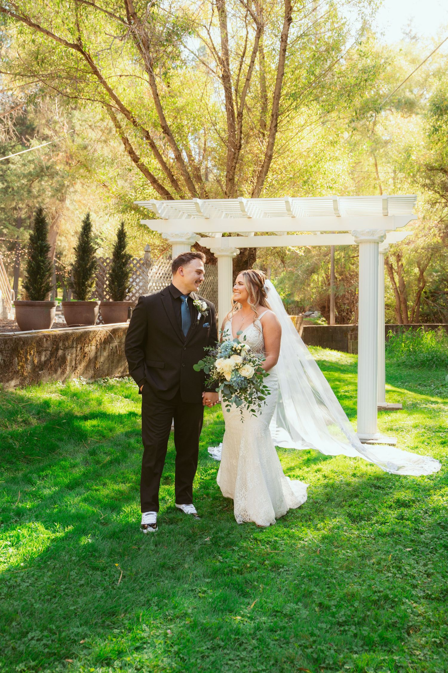A romantic wedding couple shares a tender moment in a lush garden setting beneath a white pergola on a sunny day.