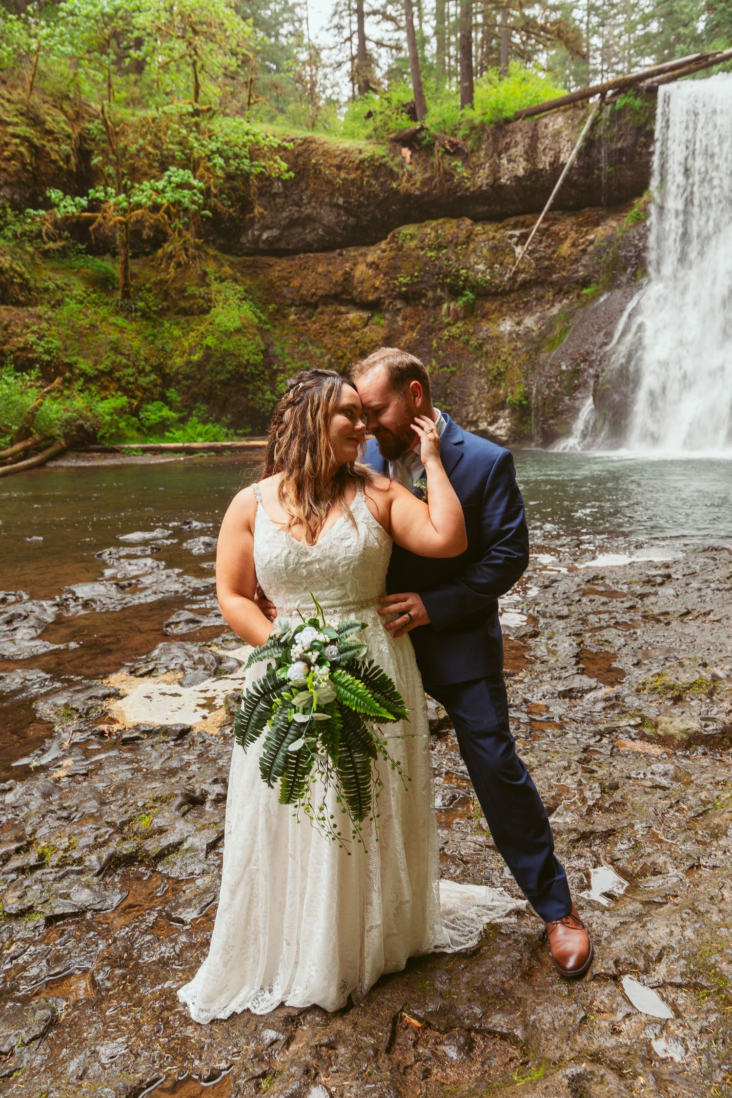 Wedding couple shares romantic moments near a forest waterfall with greenery.