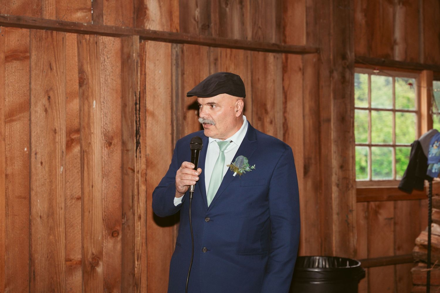 Groomsman in dark suit and cap gestures during wedding toast in wooden barn.