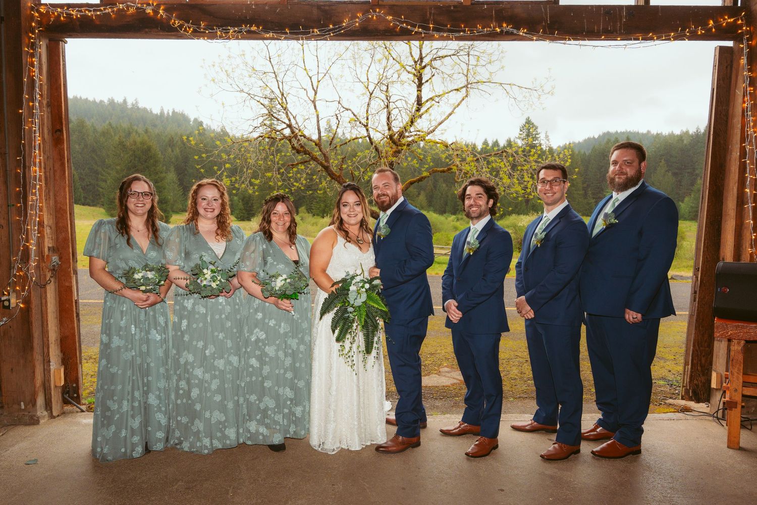 Wedding party in sage green bridesmaid dresses and navy suits pose under wooden arch in outdoor setting.