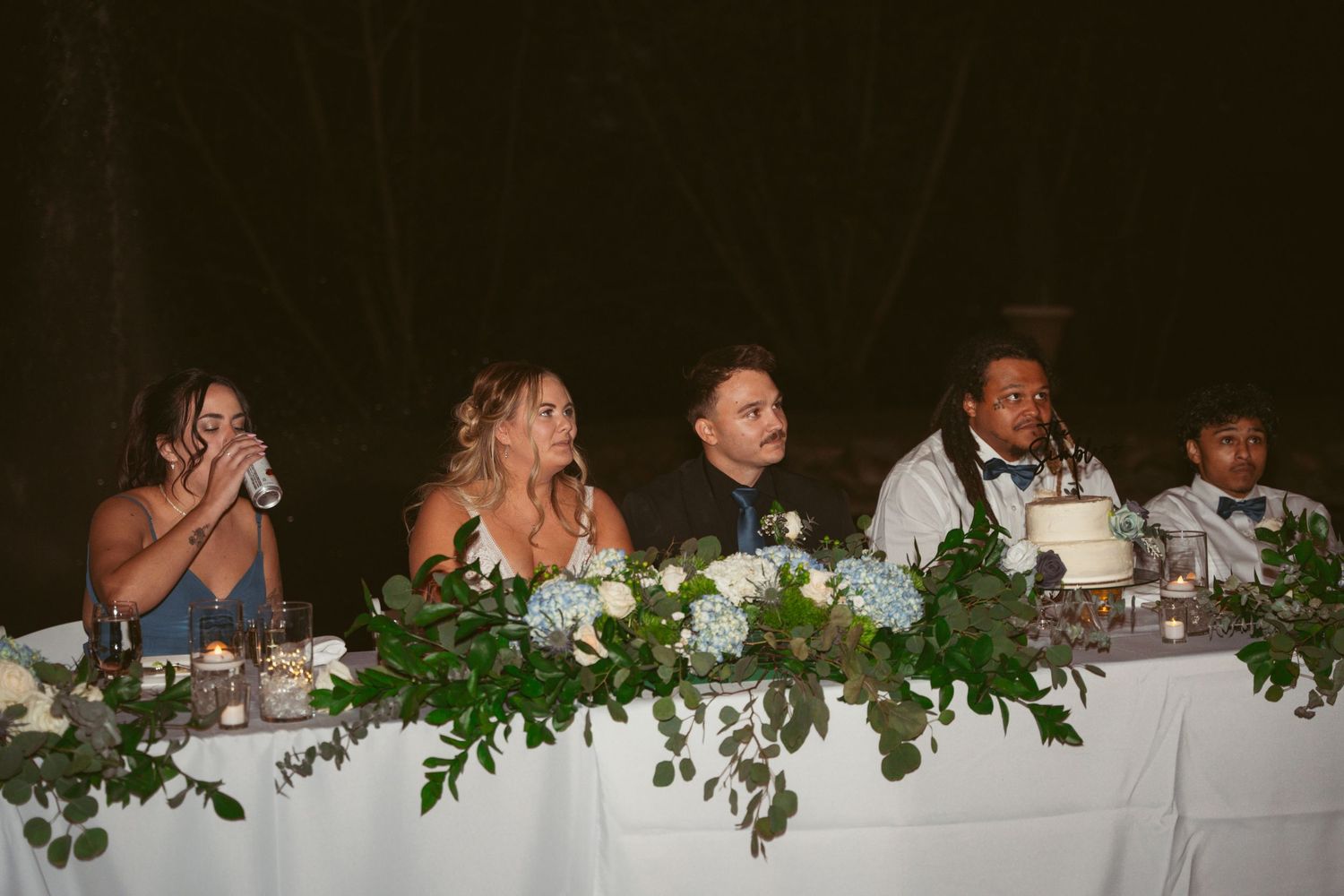 Long reception table with white tablecloths and leafy green decorations at an evening wedding.
