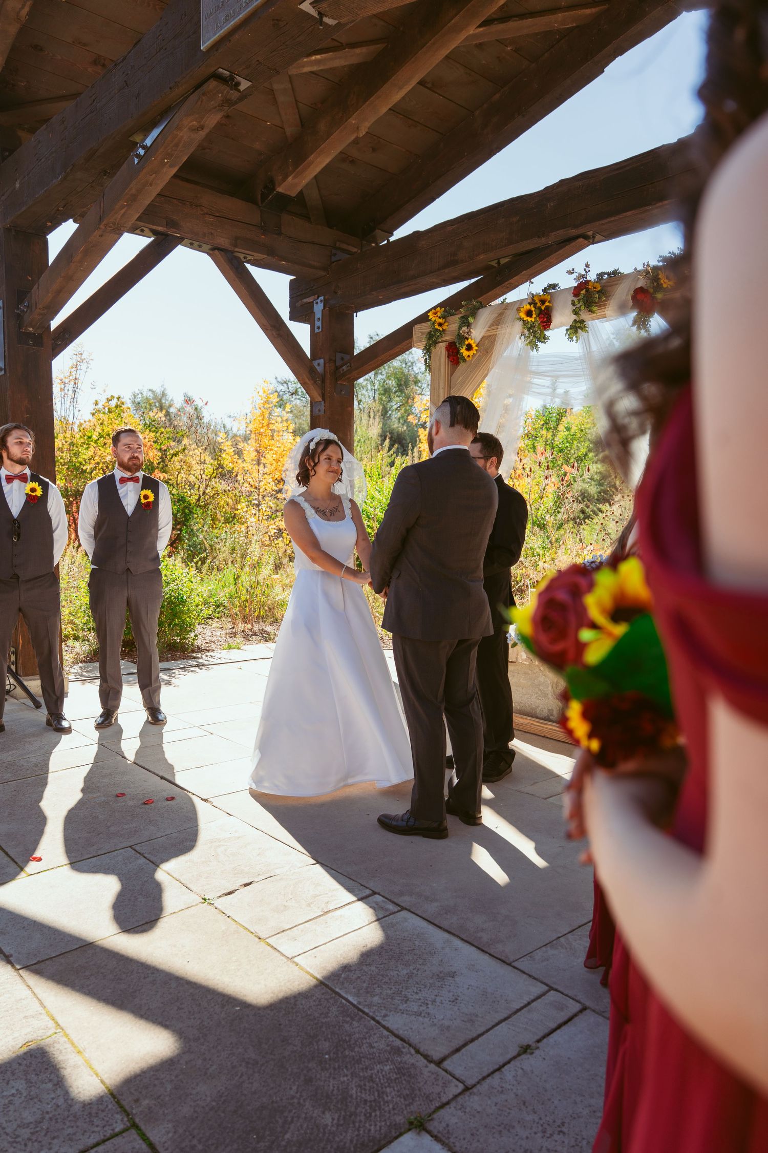 Sunlit outdoor wedding ceremony under a wooden pavilion with autumn foliage in the background.