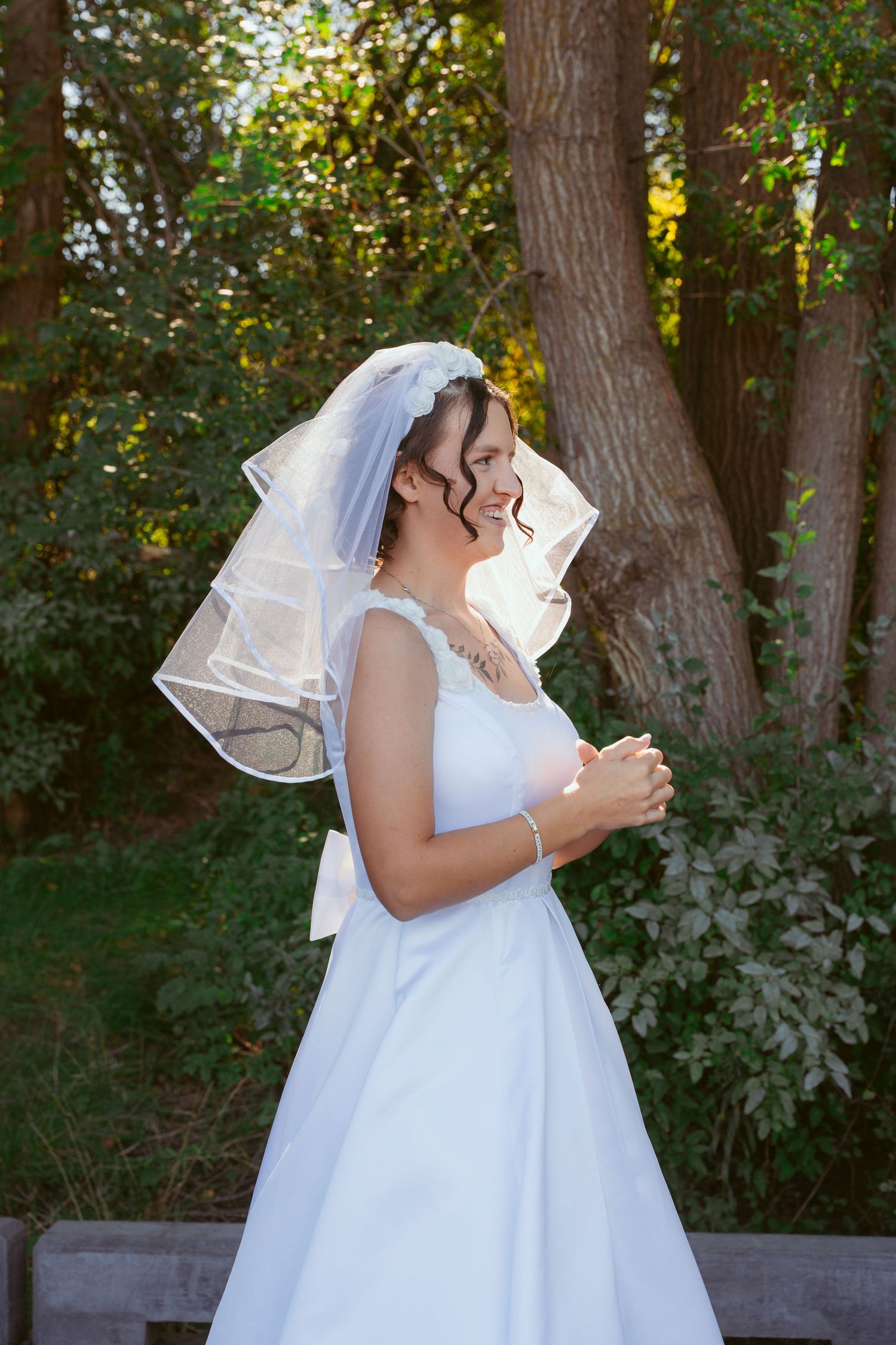 A bride in a white wedding dress and veil stands in a sunlit wooded area.