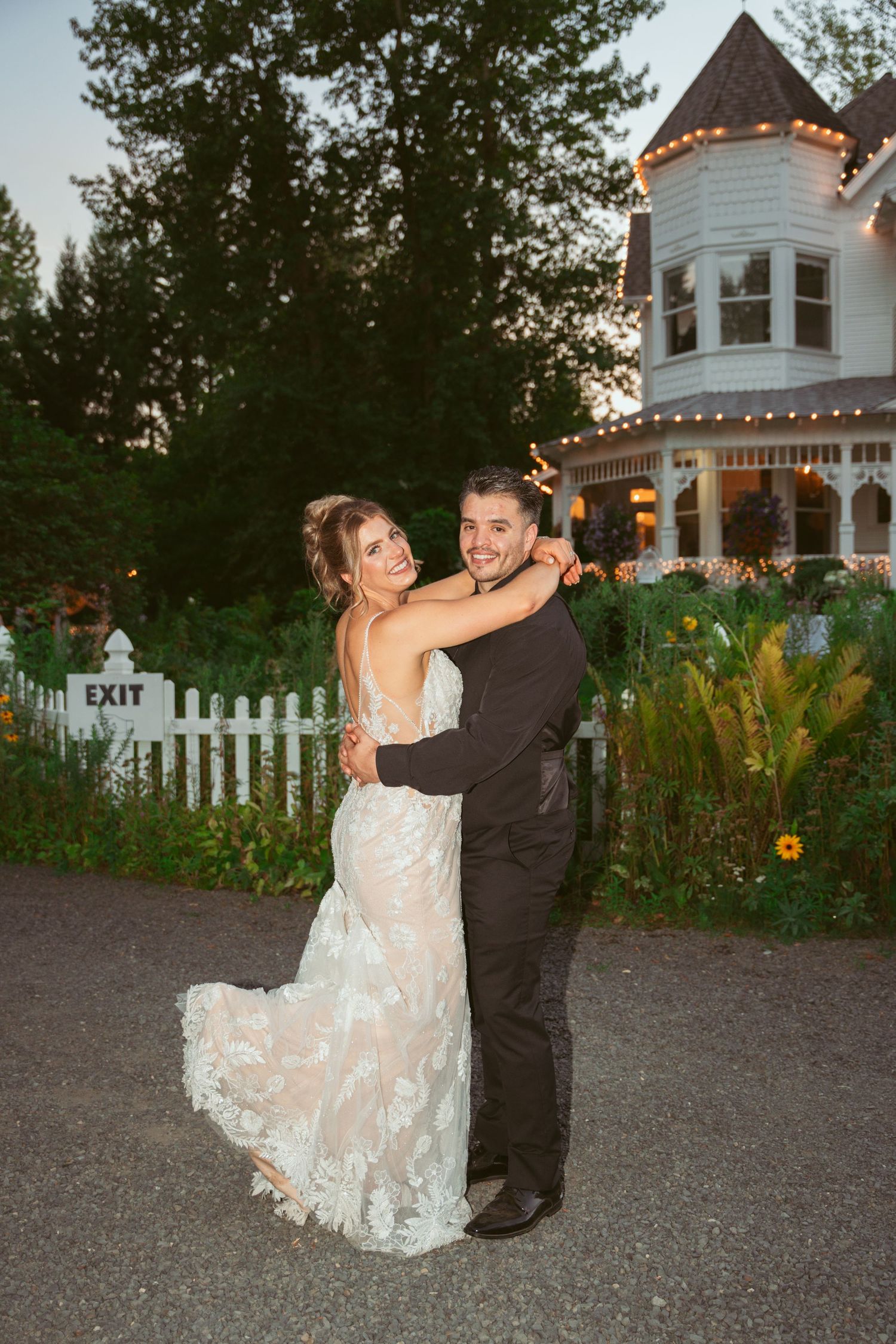 Couple dances and twirls together on gravel path near white picket fence and gazebo.