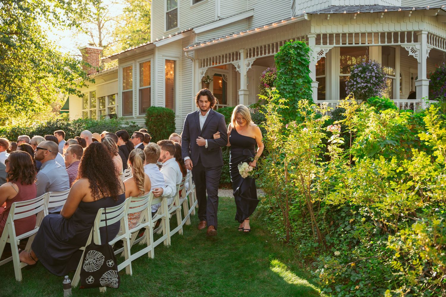Wedding guests seated on white chairs on a lawn with historic house and garden in background.