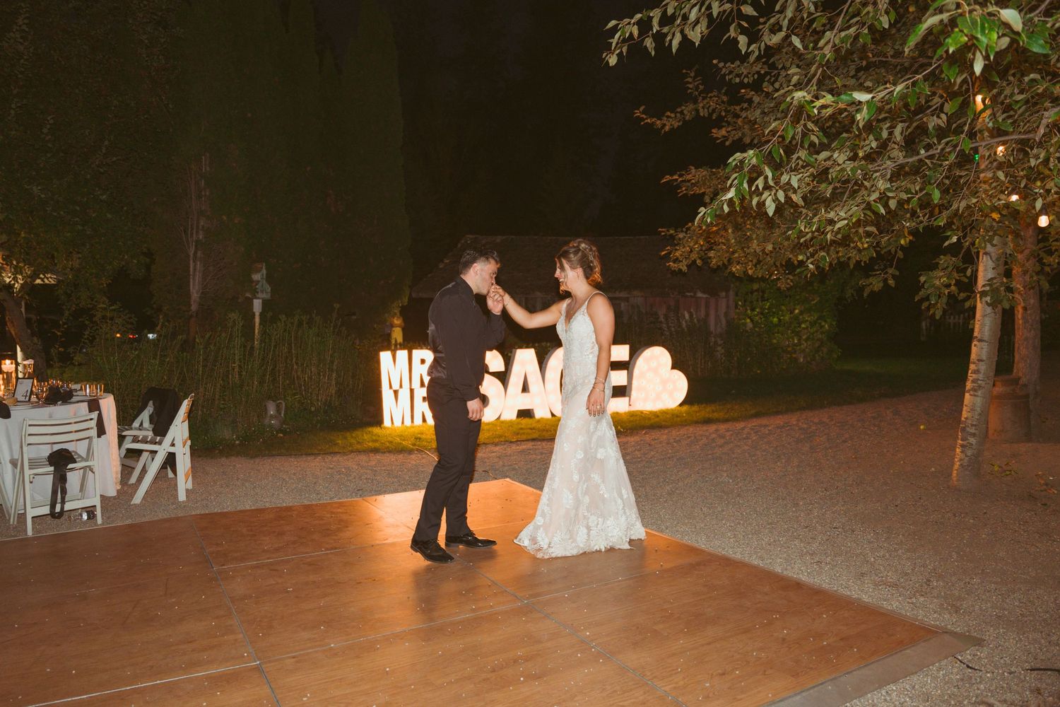 Couples dancing on an outdoor wooden dance floor at dusk surrounded by trees and string lights.