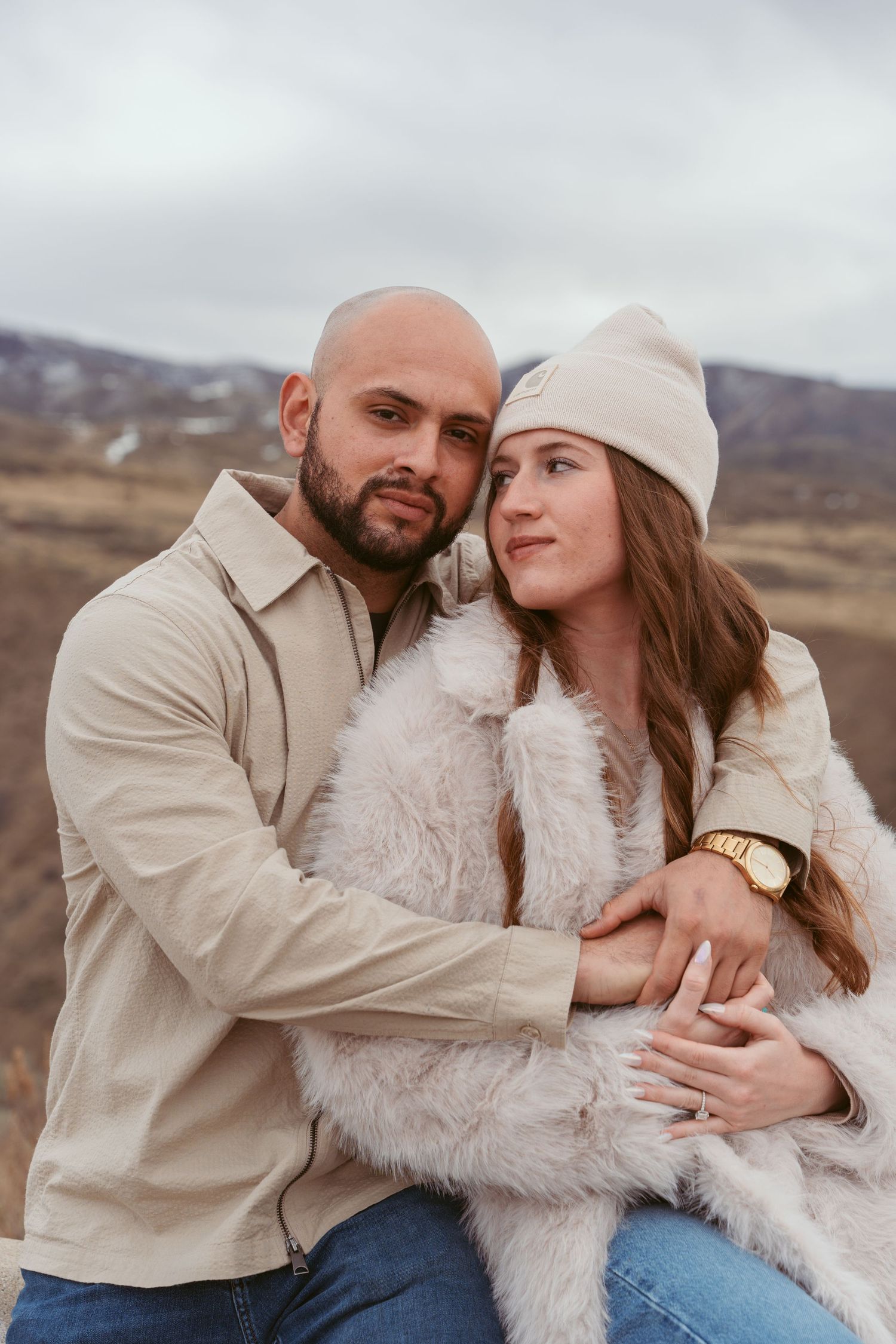 A couple embraces in warm winter clothing against a backdrop of rolling hills on an overcast day.