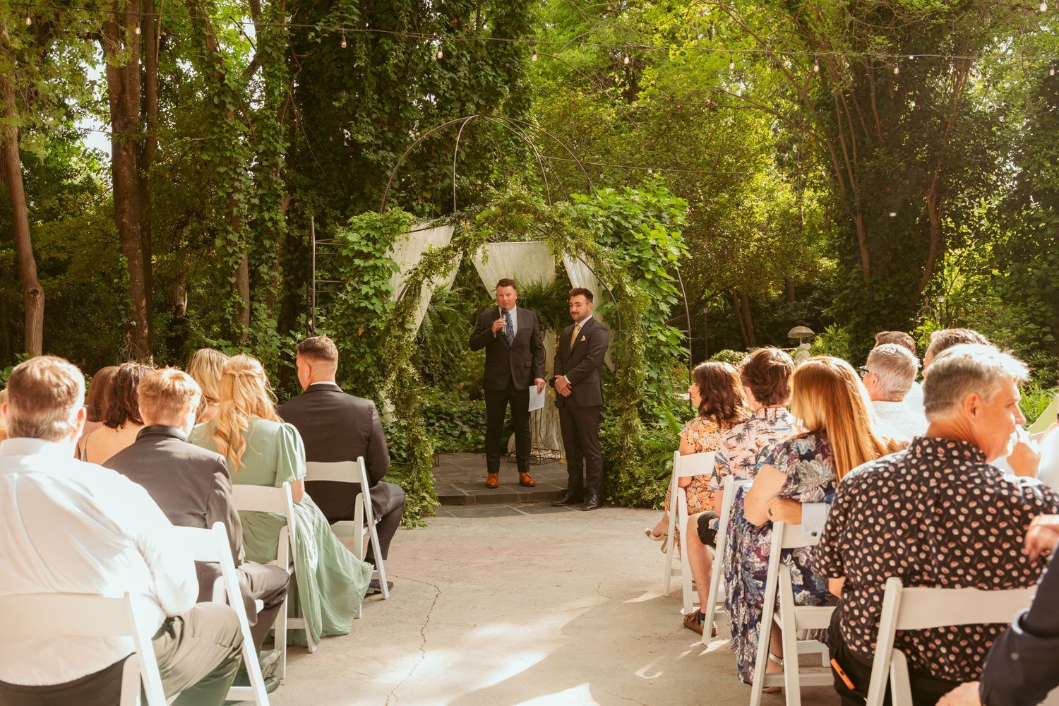 Garden wedding ceremony with white chairs and floral arch under trees.