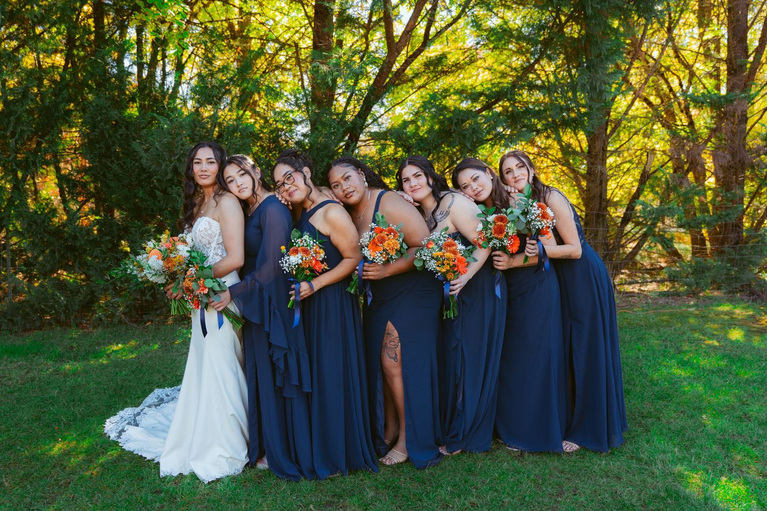 Wedding party poses casually on grass with navy blue dresses and orange floral arrangements.