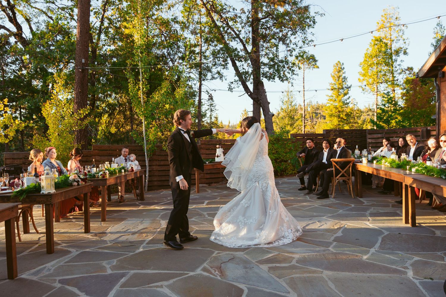 Couple dances together on rustic stone patio surrounded by pine trees during sunset celebration.