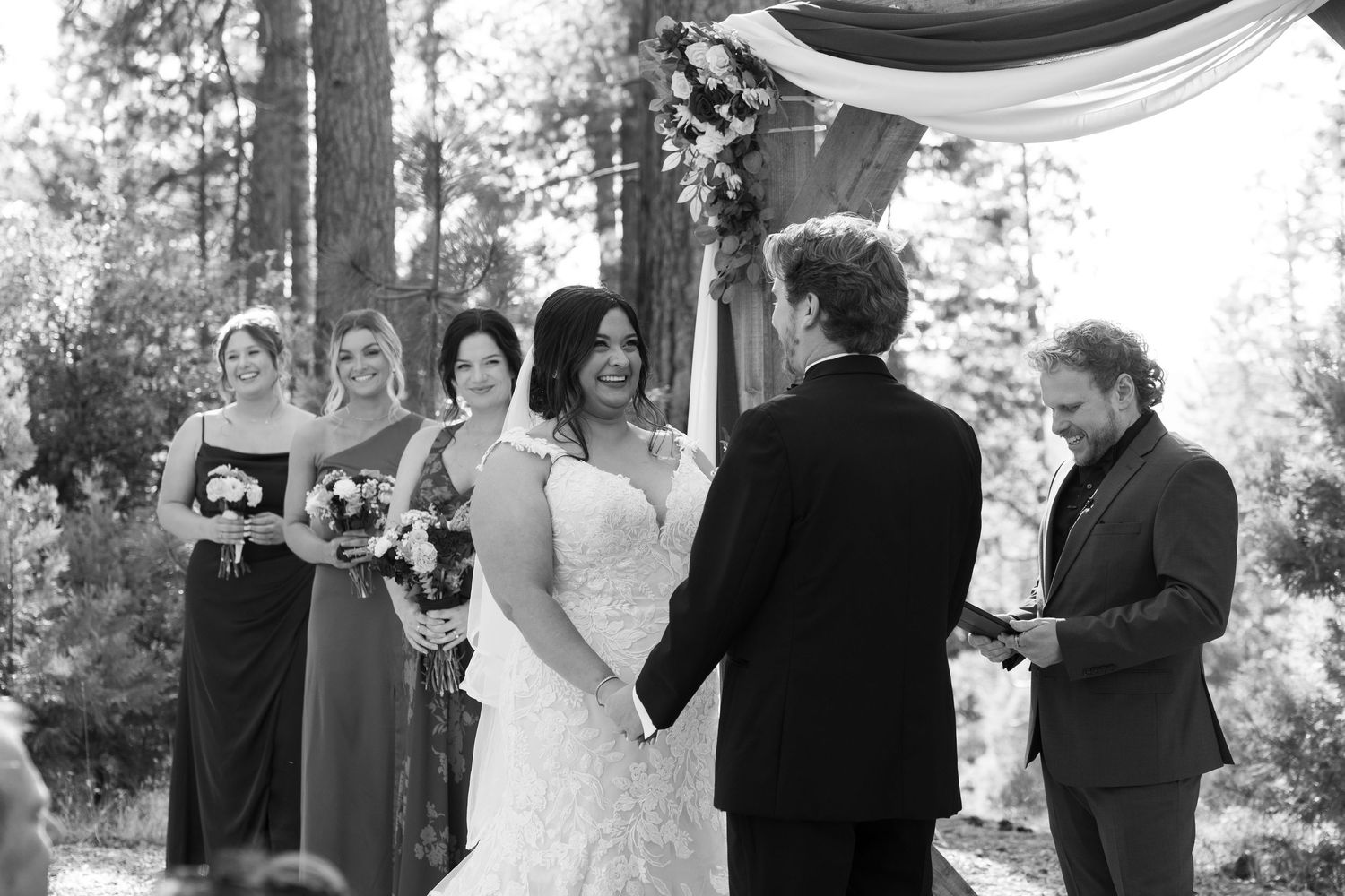 Black and white photo of wedding ceremony under wooden arch in pine forest.