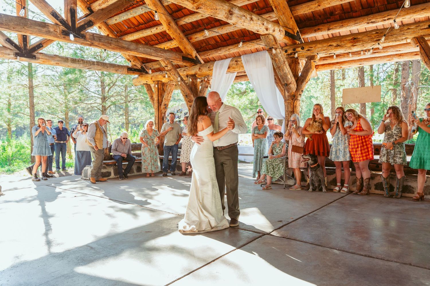 Wedding celebration in an open-air wooden pavilion surrounded by pine trees.