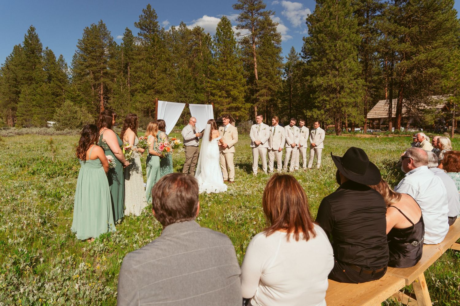 Wedding guests watch ceremony from wooden benches in a scenic mountain meadow setting.