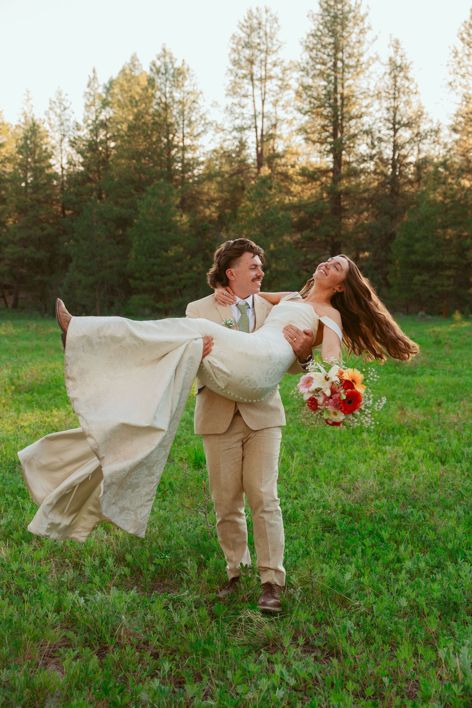Newlyweds share a playful dip pose in a green field with evergreen trees in the background.