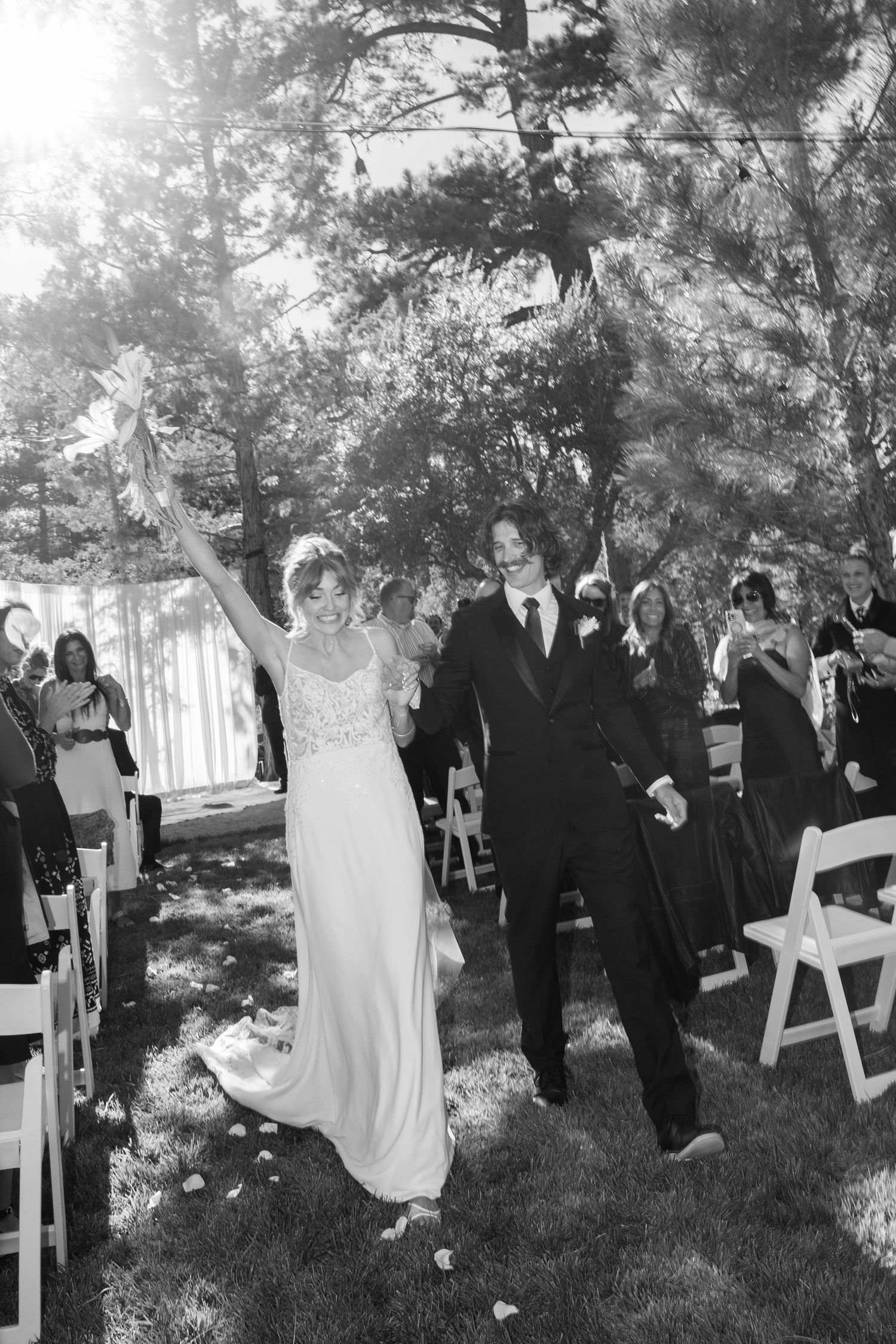 Black and white photo of wedding guests celebrating at an outdoor ceremony under pine trees.