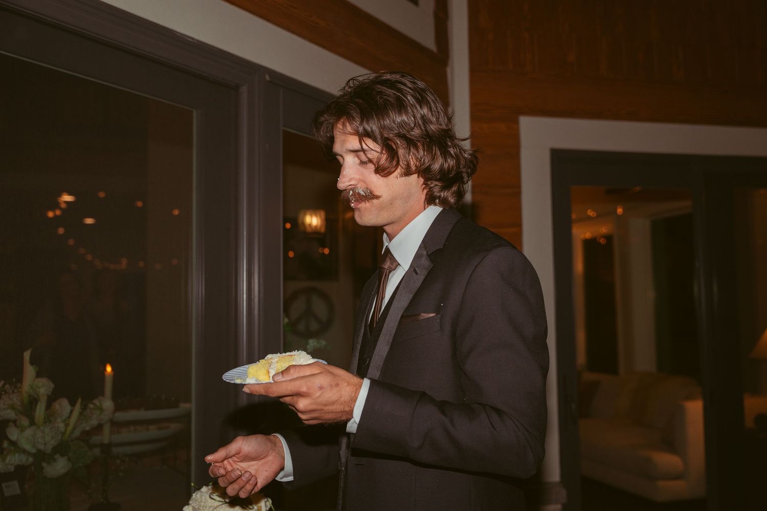 A groom in a dark suit holds a plate of wedding cake during the reception.