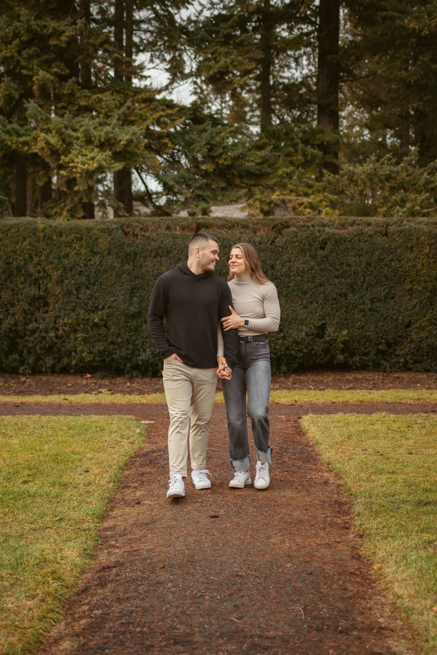 Couple walking together on a winding garden path surrounded by hedges and trees.