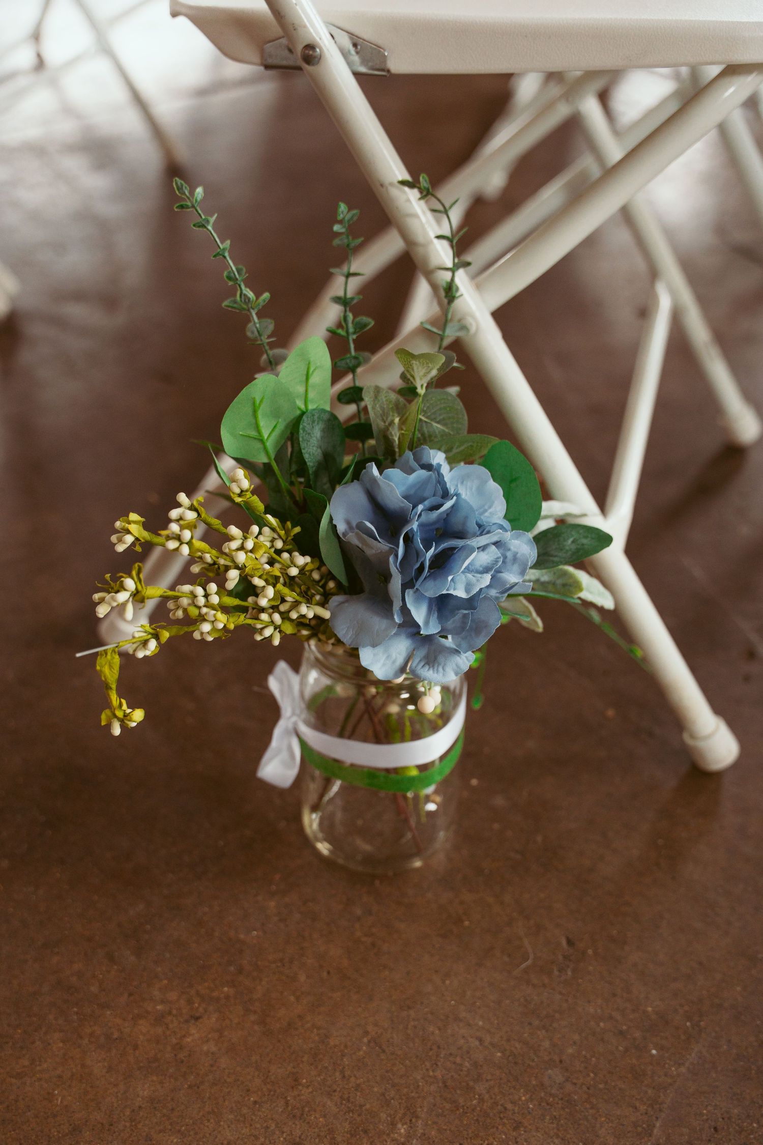 Mason jar flower arrangements with blue hydrangeas and yellow accents on wooden chairs.
