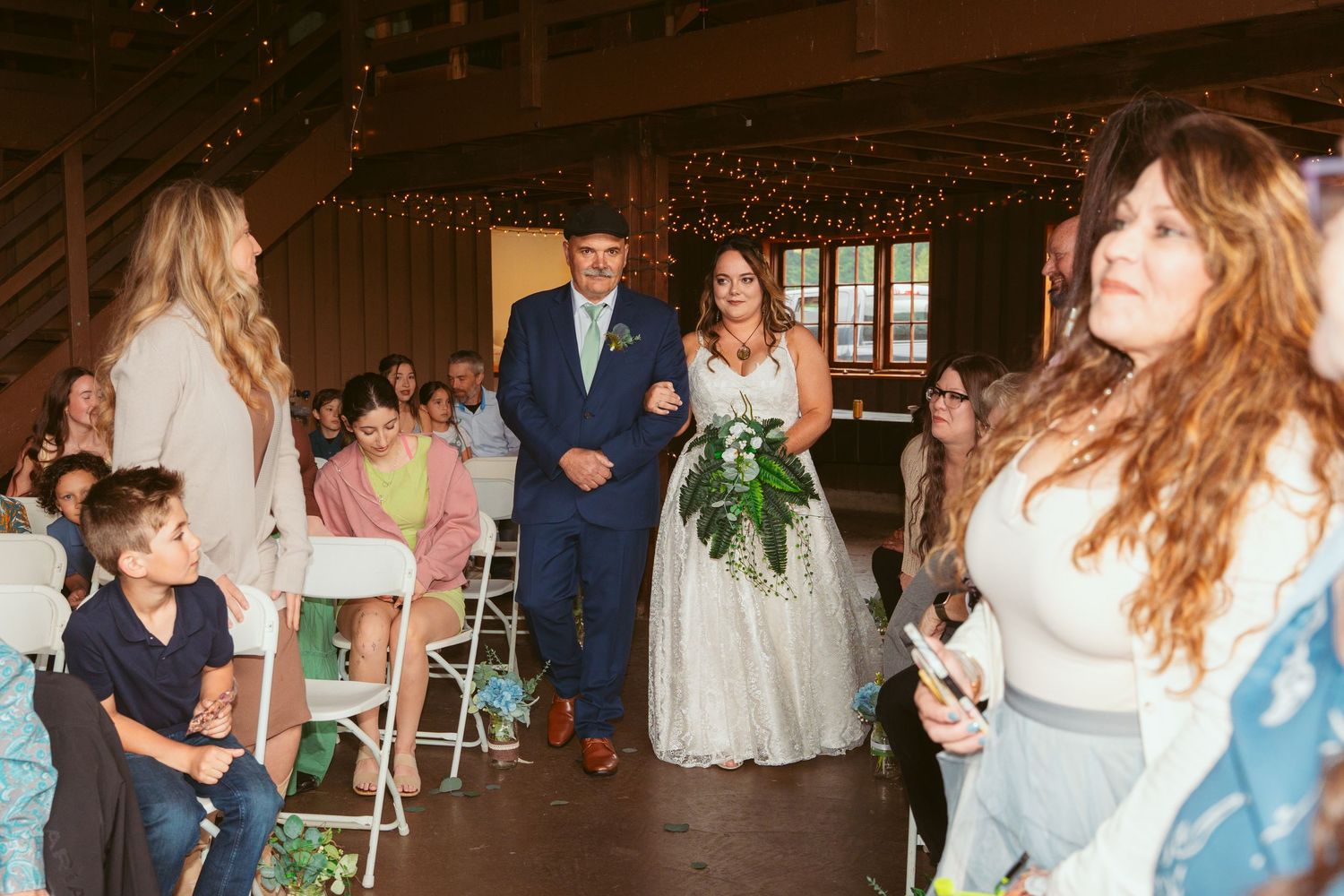 Wedding guests celebrate inside a wooden barn decorated with string lights and greenery.