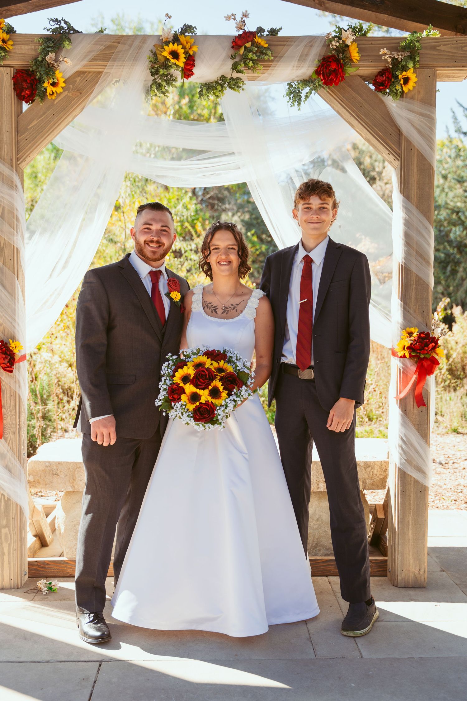 Wedding party members in burgundy and grey attire pose under decorated pavilion.