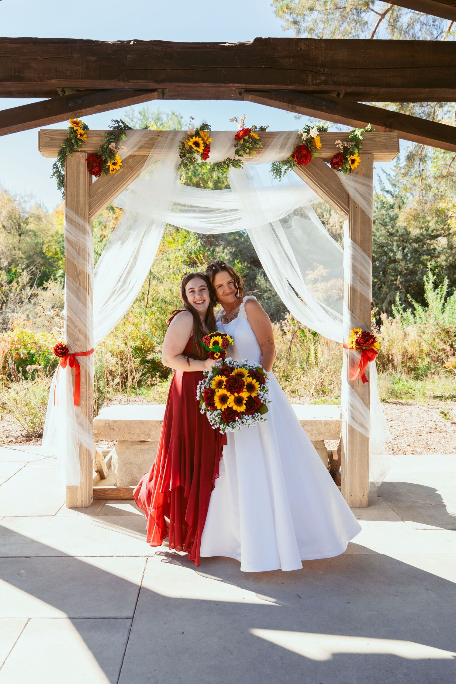 Wedding party shares candid moments under sunflower-decorated pergola with white fabric draping.