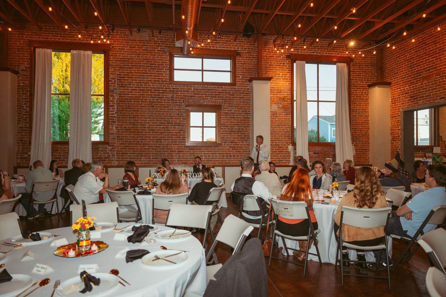 Reception hall decorated with round tables and string lights hanging from exposed brick walls.