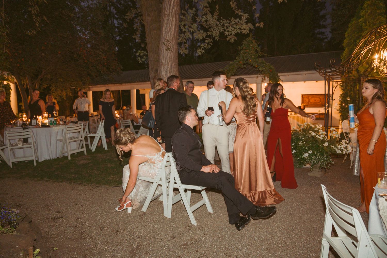 Wedding guests dance and mingle at an outdoor evening reception with string lights and rustic decor.