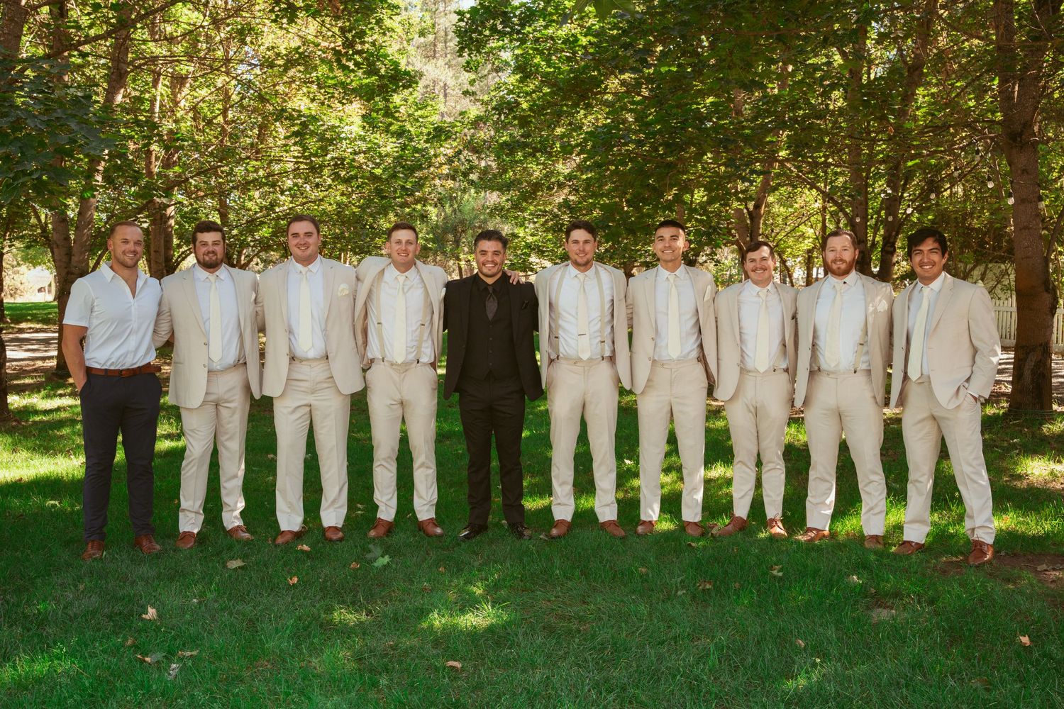 Wedding party members in cream colored suits line up for a formal photo in a garden setting.