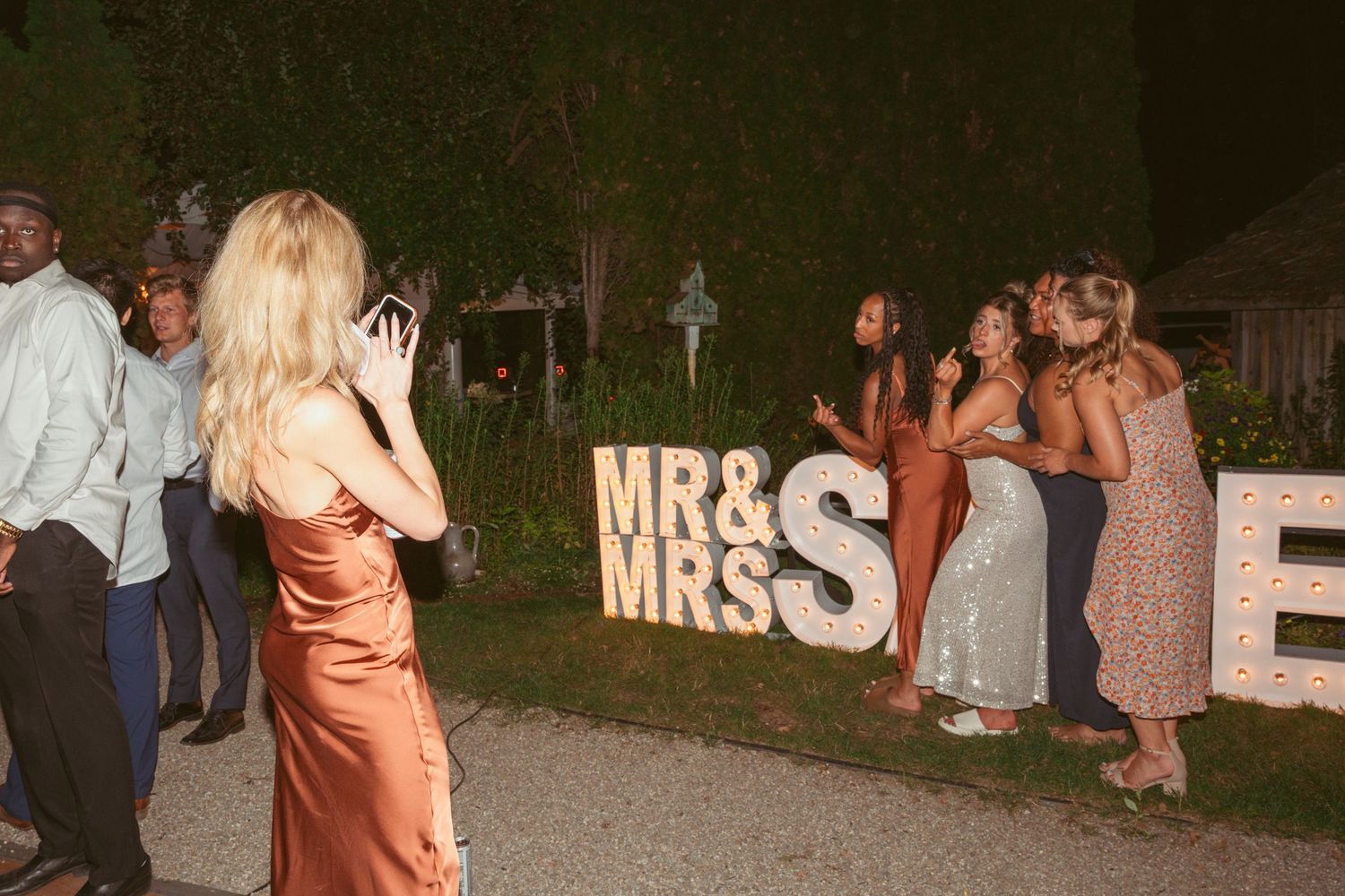 Guests taking photos by illuminated 'MR & MRS' sign at an outdoor wedding reception.