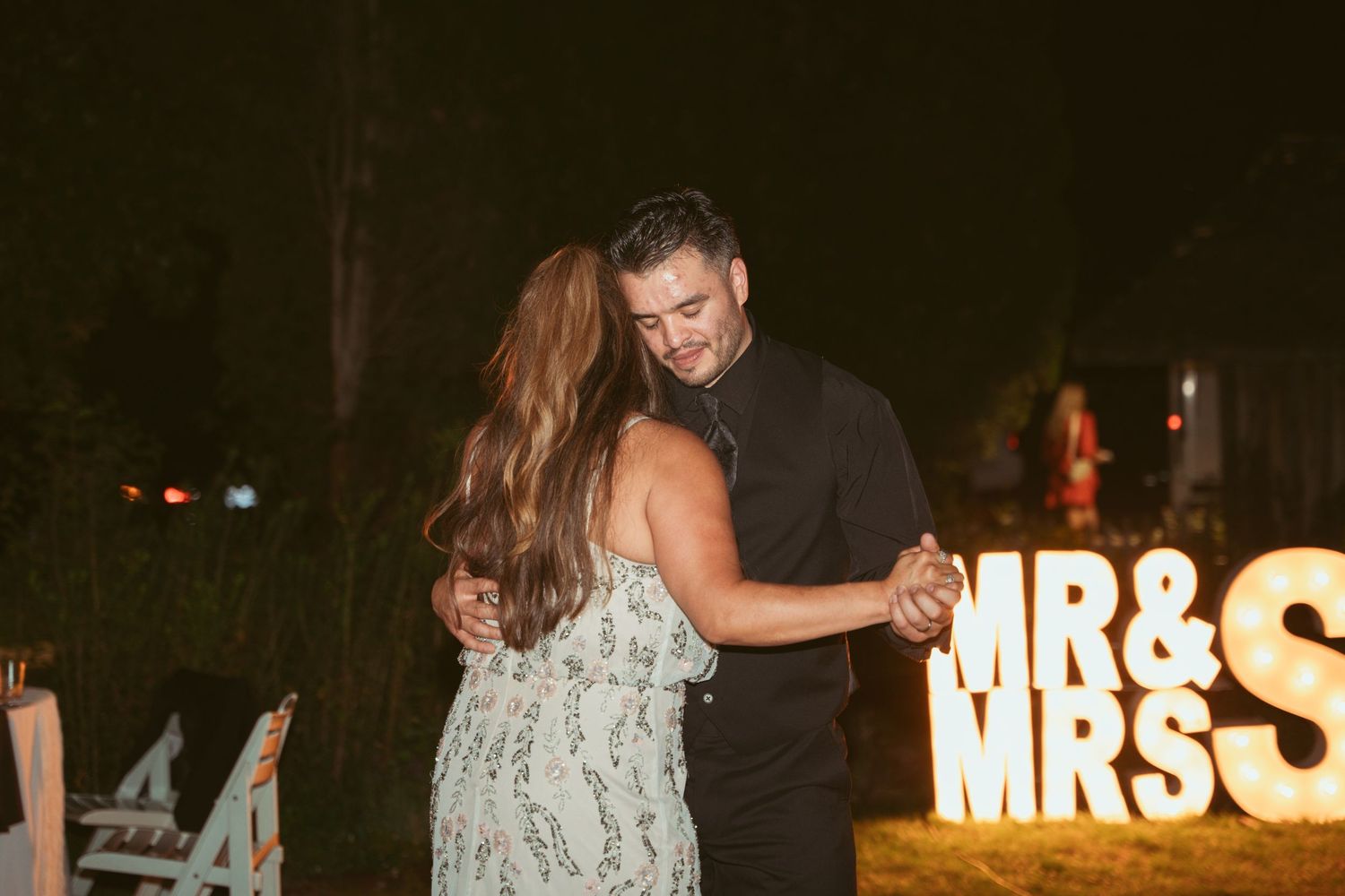 Wedding reception dance party at night with illuminated MRS sign and guests dancing.