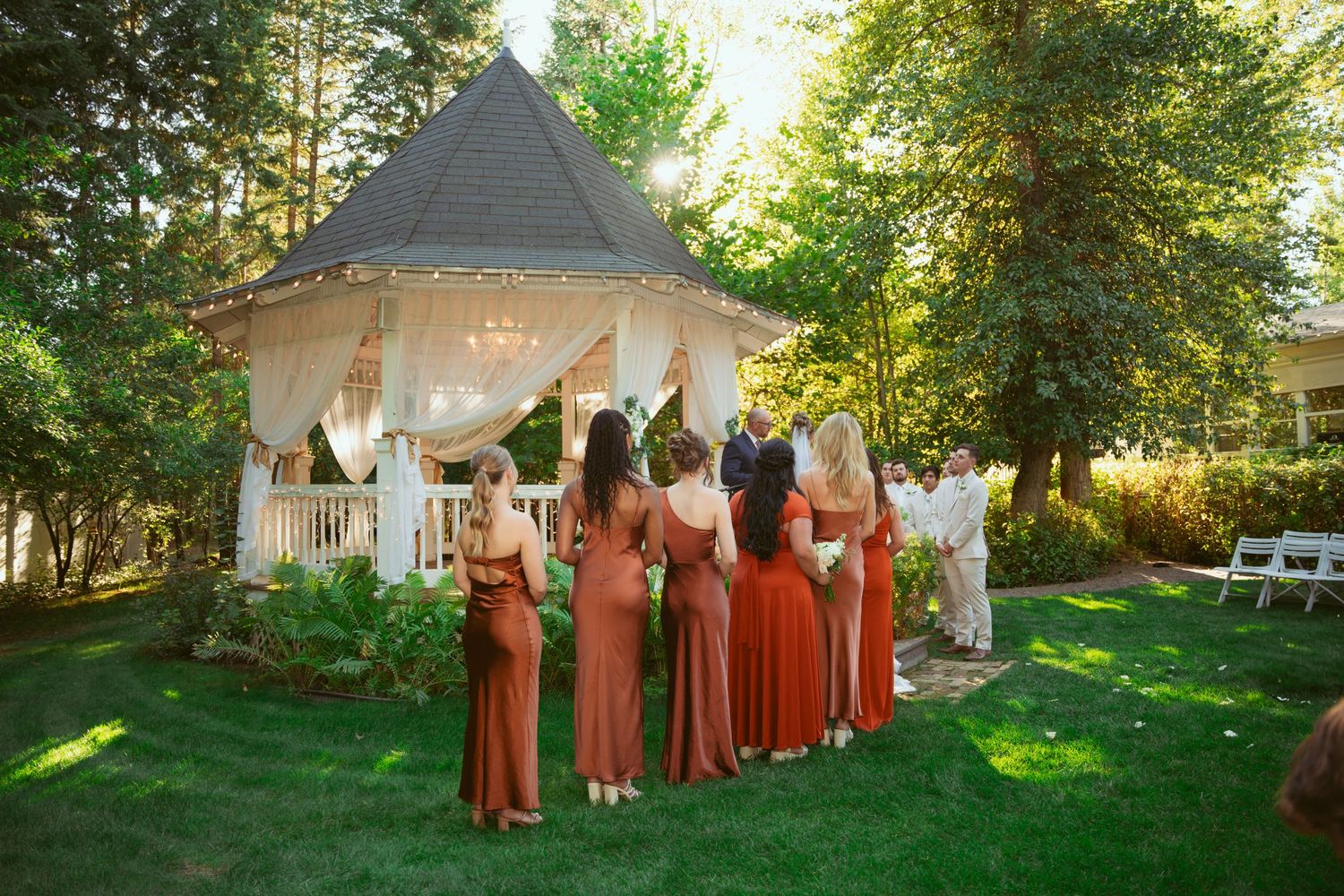 Wedding party in rust-colored dresses poses together on a sunny lawn with a white gazebo in the background.